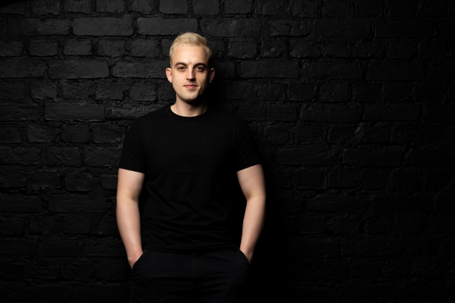 Vīrieša portrets melnā ķieģeļu sienas telpā.